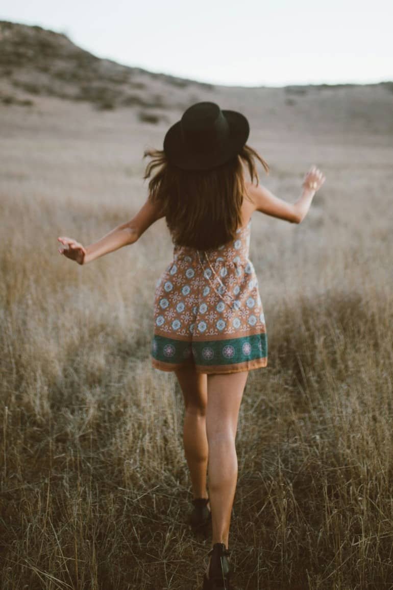 A woman wearing a patterned romper and a black hat is walking through a dry, grassy field with hills in the background.