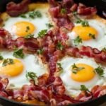 A black skillet filled with four sunny-side-up eggs topped with fresh parsley, surrounded by crispy bacon. The scene conveys a warm, appetizing breakfast setting.