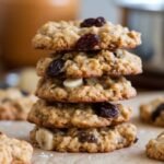 A stack of five oatmeal cookies with raisins and nuts on a parchment-lined surface. More cookies are blurred in the background. Warm and rustic tone.