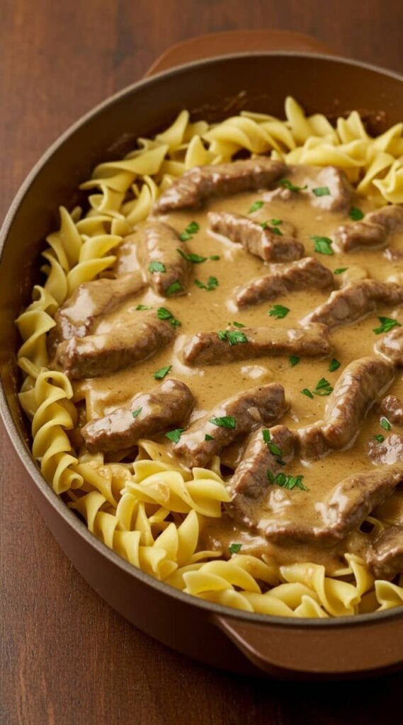 A round dish filled with creamy beef stroganoff, featuring tender brown beef strips in savory sauce, garnished with green herbs, and surrounded by curly egg noodles.