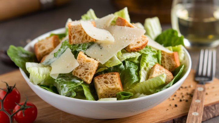 A fresh Caesar salad in a white bowl topped with croutons and shaved Parmesan cheese. A glass of white wine and cherry tomatoes sit nearby.