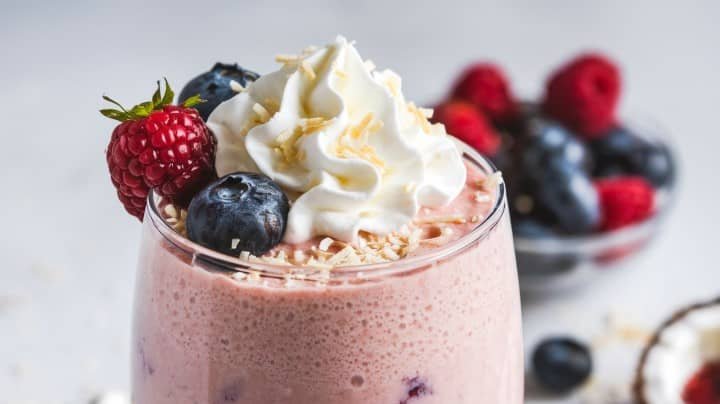 A creamy smoothie topped with whipped cream, a raspberry, a blueberry, and toasted coconut flakes. A dish of mixed berries sits in the background.