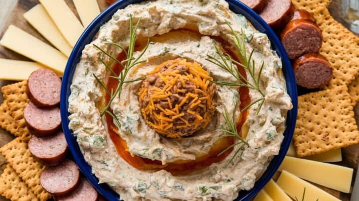 A platter with creamy dip topped with a cheese ball and garnished with rosemary. Surrounded by sliced sausage, cheese, and crackers, creating a cozy, inviting feel.