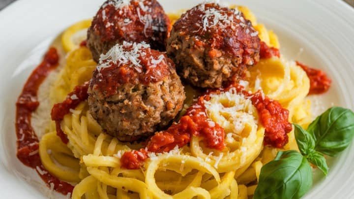 A plate of spaghetti with three large meatballs, topped with red tomato sauce and grated cheese, garnished with fresh basil leaves. The dish looks savory and appetizing.