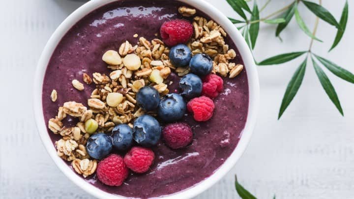 A vibrant acai bowl topped with granola, blueberries, and raspberries sits on a white surface. Green leaves frame the image, adding a fresh, natural feel.