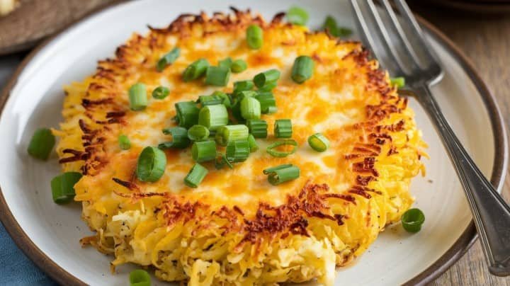 A crispy golden cauliflower hash brown on a white plate, topped with melted cheese and fresh green onions, next to a fork, creating a savory, inviting look.