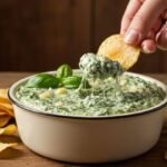 A hand dips a chip into creamy spinach dip in a white bowl, garnished with basil leaves. Chips are scattered on a rustic wooden table, creating a cozy vibe.