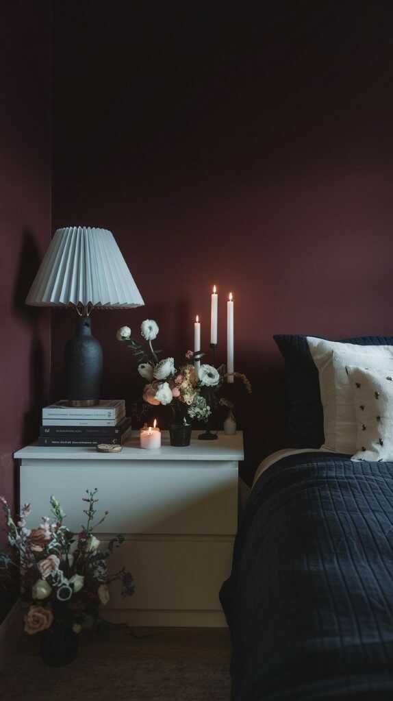 A cozy dark bedroom vignette featuring candles, books, and flower arrangements.