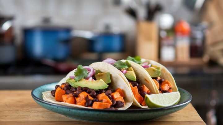 Three tacos filled with black beans, sweet potatoes, avocado, red onion, and cilantro sit on a green plate with a lime wedge, in a cozy kitchen setting.