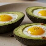 Three avocado halves on a beige plate, each cradling a baked egg with a sunny yolk. Light seasoning of salt and pepper adds a savory touch.