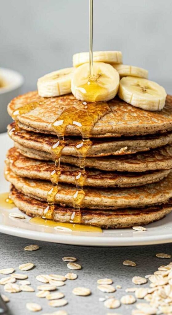 Stack of golden banana oatmeal pancakes topped with banana slices, drizzled with honey. Oats scattered on a gray table complete the cozy breakfast scene.