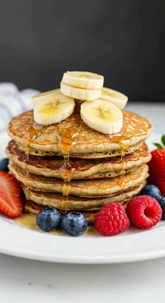 A stack of banana oatmeal pancakes topped with banana slices and drizzled with syrup. Fresh strawberries, raspberries, and blueberries surround the pancakes.