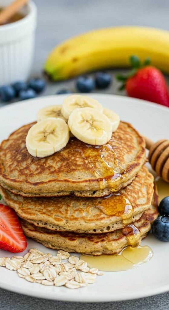 Stack of banana oatmeal pancakes topped with banana slices and honey, surrounded by strawberries, blueberries, oats, and a banana in the background. Fresh and inviting.