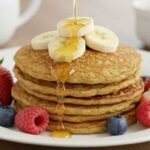 A stack of pancakes topped with banana slices is drizzled with syrup on a white plate, surrounded by strawberries, raspberries, and blueberries. A mug and bowl are in the background.