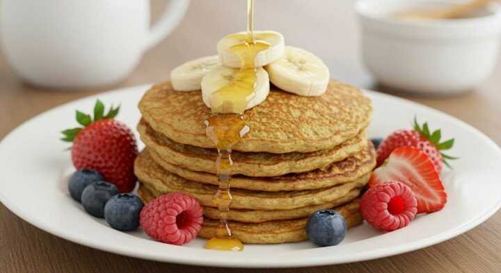 A stack of pancakes topped with banana slices is drizzled with syrup on a white plate, surrounded by strawberries, raspberries, and blueberries. A mug and bowl are in the background.