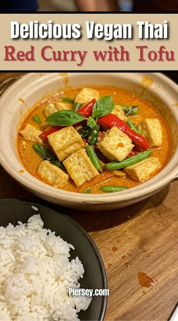 A bowl of vibrant vegan Thai red curry with tofu, green beans, red peppers, and basil. A side of white rice sits nearby on a wooden table.