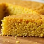 A slice of golden-brown keto cornbread on a wooden board. The texture appears moist and crumbly, evoking a warm, comforting, and homemade feel.