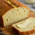 A freshly baked loaf of keto egg bread with a slice cut off, topped with a pat of melting butter, rests on a wooden cutting board. A knife lies nearby.