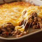 A close-up of keto hamburger casserole in a baking dish, with a wooden spoon lifting a portion. Melted cheese stretches, revealing ground beef beneath.