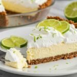 A slice of creamy key lime pie on a white plate, topped with whipped cream and lime slices. The pie has a graham cracker crust; whole limes are in the background.