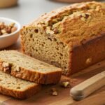 A freshly baked loaf of nut bread on a wooden cutting board, surrounded by walnuts and a knife. Two slices are cut, revealing a soft, nutty texture.