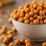 A bowl filled with roasted, spiced chickpeas sits on a table. The scattered chickpeas and warm, golden hues create a cozy, appetizing atmosphere.