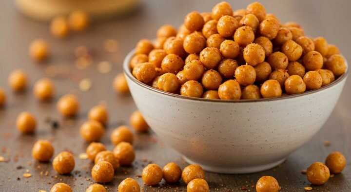 A bowl filled with roasted, spiced chickpeas sits on a table. The scattered chickpeas and warm, golden hues create a cozy, appetizing atmosphere.