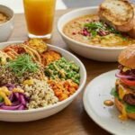 A colorful array of dishes on a wooden table, featuring a vibrant grain bowl, a hearty burger with toppings, two soups with bread, and a glass of orange juice.