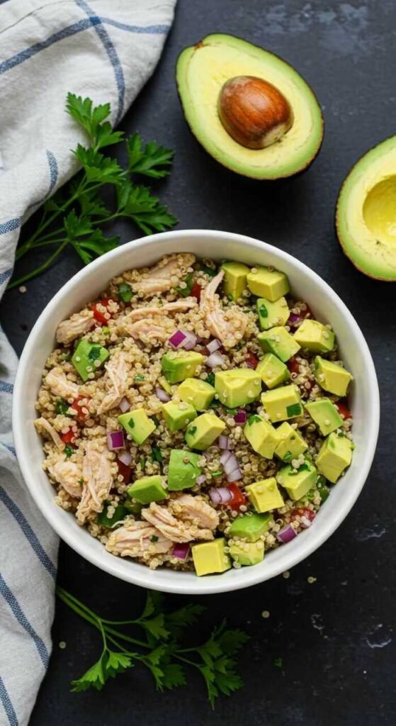 A vibrant quinoa salad with diced avocado, shredded chicken, red onion, and herbs in a white bowl. Fresh avocado halves and parsley garnish nearby.