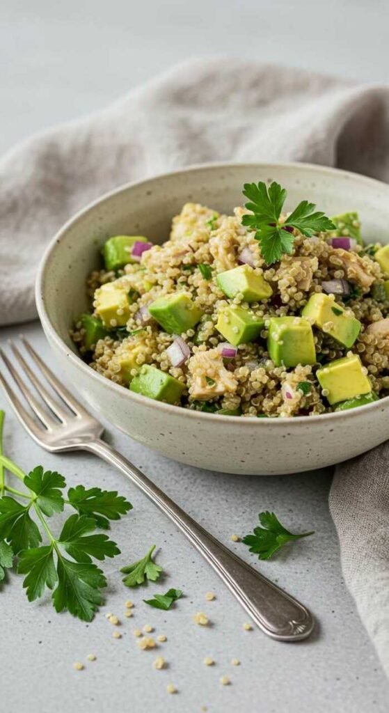 A bowl of quinoa salad with avocado chunks, red onion, and parsley garnish on a grey cloth. A fork and fresh parsley are on the side, creating a fresh and healthy vibe.