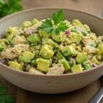 A bowl of quinoa salad with avocado, shredded chicken, and red onion, garnished with parsley. A fork and napkin are beside it on a wooden table.