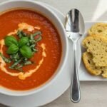 A bowl of vibrant red tomato soup garnished with cream and fresh basil sits on a wooden table. Beside it, slices of bread and a spoon, in soft daylight.
