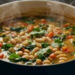 Steaming pot of bean soup in a blue pot with white beans, chopped tomatoes, and fresh green herbs. Cozy and inviting atmosphere, perfect for comfort food.