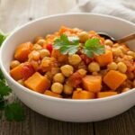 A bowl of vibrant chickpea and sweet potato stew garnished with cilantro on a rustic wooden table. Bright, hearty, and inviting.