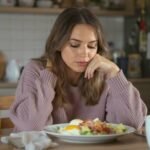 Quitting Keto & Carnivore: What 9 Years Taught Me Woman in a cozy kitchen, wearing a lavender sweater, gazes thoughtfully at a breakfast plate with eggs and bacon. Mood is contemplative and serene.