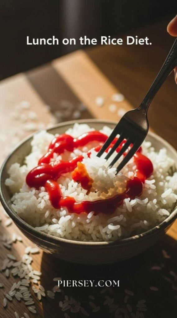 A bowl of white rice with ketchup drizzled on top is on a wooden surface. A fork is picking up a bite. Caption reads "Lunch on the Rice Diet."