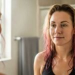 Beating Food Cravings Without Breaking Your Diet A woman with wet, wavy hair and a hint of a smile looks at her reflection in the bathroom mirror. The setting is well-lit and calm, with a towel hanging in the background.